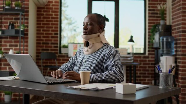 African american businessman wearing medical cervical neck collar foam after physical injury and accident. Working with pain, illness and health problem, tired stressed hurt freelancer in office.