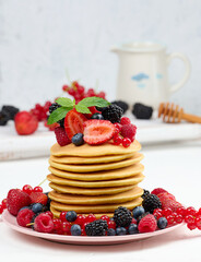 Stack of baked pancakes with fruits in a round plate on a white table