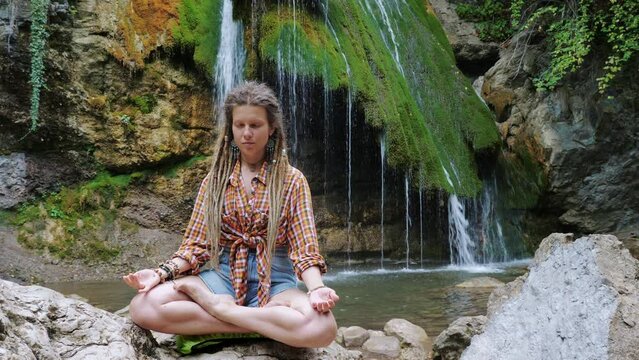 Girl with dreadlocks and boho jewelry sits in lotus position on stone near waterfall. Young woman practicing yoga, padmasana on mount. Yogini spends time in meditation, contemplation in wildland