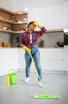 Tired sad young black lady housewife in rubber gloves with mop washes floor, wipes forehead with hand