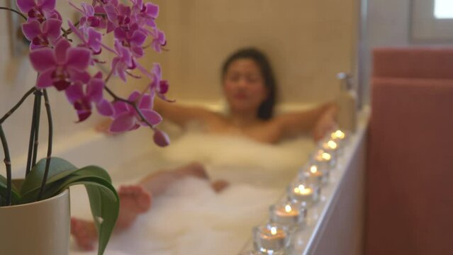 Purple blooming orchid with lined up glowing candles for creating relaxing atmosphere. Tranquil ambience for wellness treatment experience. Beautiful young woman enjoying and relaxing in bubbly bath.