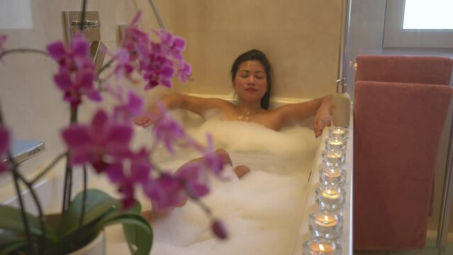 Beautiful flowering orchid with pretty young lady relaxing in nice bubble bath. Beautiful young woman enjoying and relaxing in bathtub. Tranquil and pleasant atmosphere for spa treatment experience.