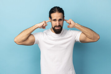 Fototapeta na wymiar I don't want to here this. Portrait of man with beard wearing white T-shirt standing and holding fingers on his ear, unpleasant sounds. Indoor studio shot isolated on blue background.