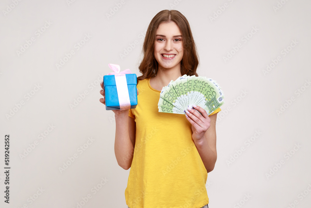 Poster Gift shopping! Happy teenager girl in yellow T-shirt holding present box and euro money banknotes, satisfied with purchase, cashback and bank loan. Indoor studio shot isolated on gray background.