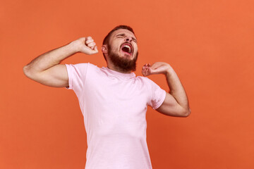 Fototapeta na wymiar Portrait of tired bearded man yawning and covering mouth with hand, feeling exhausted, lack of sleep, outstretching arms, wearing pink T-shirt. Indoor studio shot isolated on orange background.