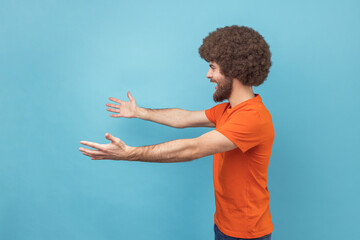 Come into my arms. Side view of satisfied man with Afro hairstyle wearing orange T-shirt reaching out to camera, stretching arms to hug you. Indoor studio shot isolated on blue background.