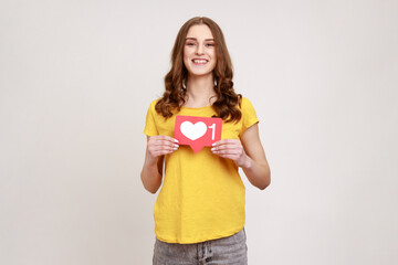 Like button of social media. Portrait of pleasant looking young woman with wavy hair holding heart icon, recommending to follow and share blog. Indoor studio shot isolated on gray background.