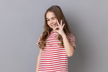 Portrait of playful adorable little girl wearing striped T-shirt showing ok sign to camera, approving something with positive expression. Indoor studio shot isolated on gray background.