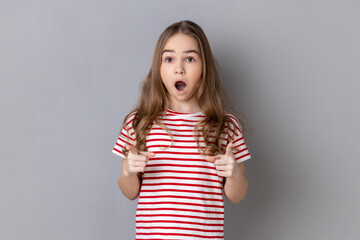Portrait of astonished little girl wearing striped T-shirt pointing to camera with surprised shocked expression and open mouth, making choice. Indoor studio shot isolated on gray background.