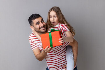 Portrait of pleased father holding his surprised daughter on his back and giving her a present for...