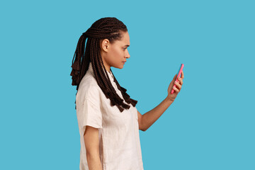 Naklejka premium Side view of woman with dreadlocks reads message on modern mobile phone, surfes social media, has concentrated look in display, updates information. Indoor studio shot isolated on blue background.