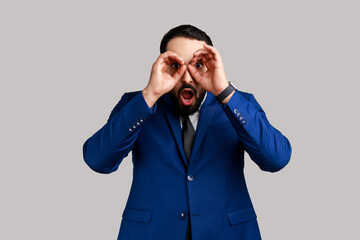 Portrait of astonished surprised bearded man standing raised arms and looking with binoculars gesture, wearing official style suit. Indoor studio shot isolated on gray background. - obrazy, fototapety, plakaty