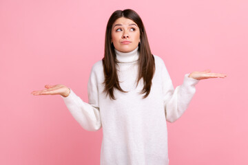 Ambiguous confused dark haired female standing with raised arms, showing helpless gesture, no answer, wearing white casual style sweater. Indoor studio shot isolated on pink background.