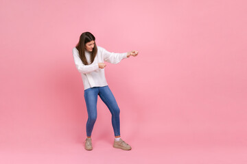 Fototapeta na wymiar Full length portrait of girl pulling invisible rope with effort, using all strength to achieve goal, wearing white casual style sweater. Indoor studio shot isolated on pink background.
