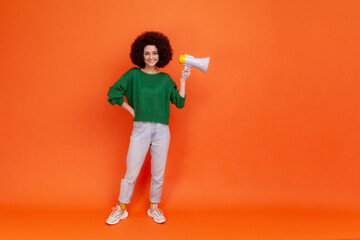 Full length photo of happy smiling woman with Afro hairstyle wearing green casual style sweater holding megaphone in hands, copy space. Indoor studio shot isolated on orange background.