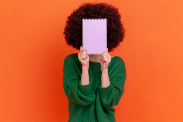 Portrait of unknown woman with Afro hairstyle wearing green casual style sweater standing holding organizer, hiding her face behind paper notebook. Indoor studio shot isolated on orange background.