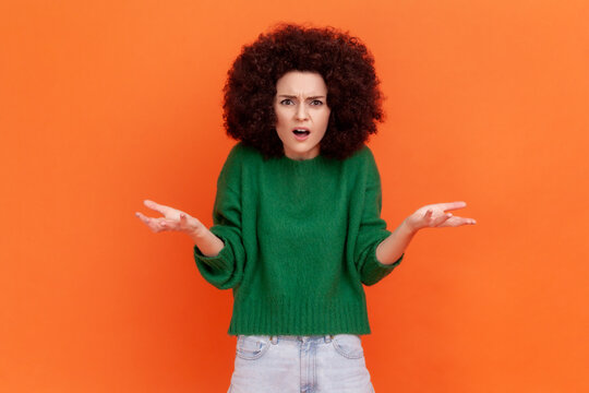 What do you want? Woman with Afro hairstyle wearing green casual sweater asking who why make this conflict, looking with annoyed indignant expression. Indoor studio shot isolated on orange background.