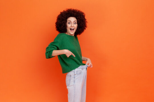 Happy woman with Afro hairstyle wearing green casual style sweater showing slim waist in big trousers, successful weight loss, diet conception. Indoor studio shot isolated on orange background.