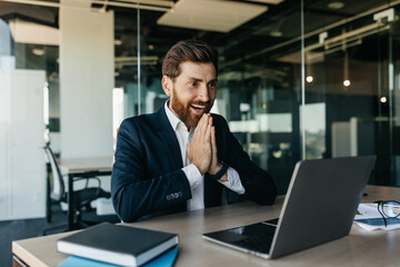 Excited businessman celebrating great news and luck, looking at laptop screen in joy, sitting in office interior