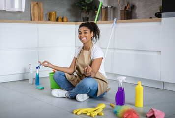 Happy calm young african american housewife sit on floor with cleaning supplies, meditating