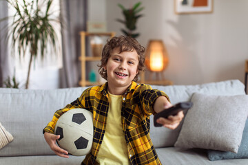 Little boy football fan watching tv at home, sitting on sofa with remote controller and soccer ball, smiling to camera