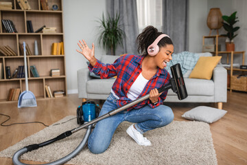 Glad satisfied millennial african american housewife in headphones vacuums and sings song to vacuum...