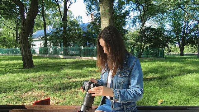 A beautiful young girl is sitting on a bench in the park looking at pictures on her camera