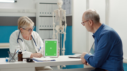 Obraz premium General practitioner vertically holding tablet with greenscreen on display in medical cabinet. Mockup template with blank chroma key background and isolated copyspace. Tripod shot.