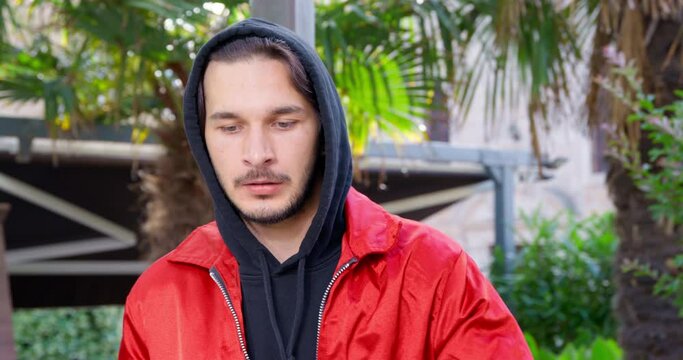Suspicious-looking young man in hoodie is stirring up something illegal, so he looks around nervously, front view. Guy rubs his nose like drug addict after another snuffed dose