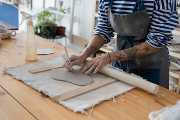 Craft artist making object out of clay in studio, cropped shot. Female ceramic artist with tattoo on hands use fettling knife to cut and trim clay, creating handmade kitchenware at pottery workshop