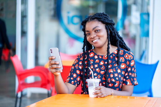 image of beautiful african lady with smart phone and a cup of drink- seated black woman having fun in a cafeteria