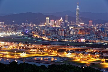 Night skyline of Downtown Taipei, the capital city of Taiwan, with 101 Tower among skyscrapers in XinYi District, a riverside park along Keelung River and lights of Songshan Airport in blue twilight