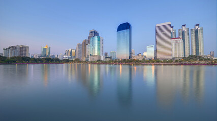Fototapeta na wymiar Night skyline of modern lakeside skyscrapers with glass curtain walls and dazzling city lights reflected in the smooth lake water in beautiful Benjakiti Park at blue dusk, in Bangkok, Thailand, Asia