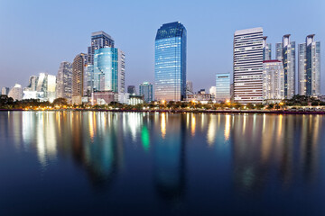 Fototapeta na wymiar Night skyline of modern lakeside skyscrapers with glass curtain walls and dazzling city lights reflected in the smooth lake water in beautiful Benjakiti Park at blue dusk, in Bangkok, Thailand, Asia
