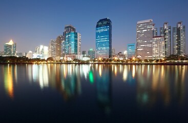 Obraz na płótnie Canvas Night skyline of modern lakeside skyscrapers with glass curtain walls and dazzling city lights reflected in the smooth lake water in beautiful Benjakiti Park at blue dusk, in Bangkok, Thailand, Asia