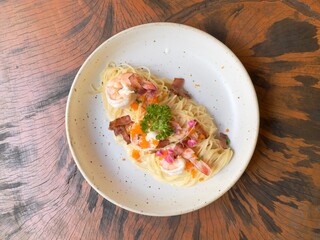 Spaghetti carbonara cream sauce with prawn, shrimp roe and crispy bacon decorated with edible flowers and parsley, served in white ceramic dish on wooden table. Selective focus