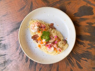 Spaghetti carbonara cream sauce with prawn, shrimp roe and crispy bacon decorated with edible flowers and parsley, served in white ceramic dish on wooden table. Top view