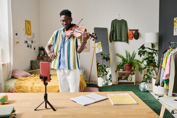 African young blogger standing in front of smartphone on tripod and playing violin for his...