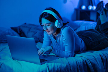 Teenage girl in wireless headphones lying on her bed and typing on laptop in her dark bedroom, she...