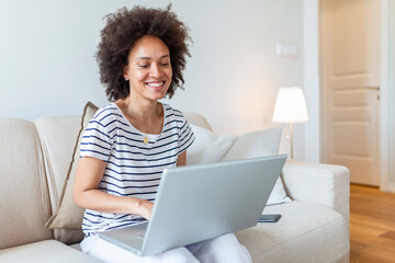 Young beautiful woman with curly hair working on laptop computer while sitting on the sofa at home
