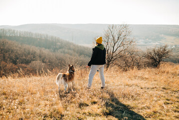 Young woman and her husky dog trek down a forest trail offering a scenic view of the trees changing...