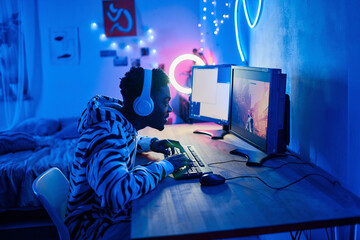 African gamer boy in wireless headphones typing on keyboard while sitting at table in front of computer and playing video game