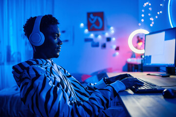 Arican teenage boy in wireless headphones playing video game on computer while sitting at table in...