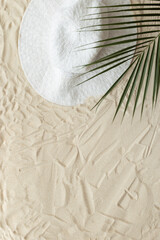 Summer minimal background, top view wide-brimmed sun hat under palm leaf on natural sand beach. Summer aesthetic photography pastel beige colored, copy space, vacation, relaxation concept
