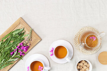 Obraz na płótnie Canvas Fireweed tea in white cups and transparent glass teapot, herbal hot tea from green leaves of ivan chai on textile tablecloth. Top view healthy drink and wild flowering willow-herb, tea time