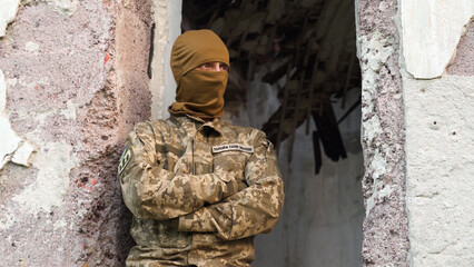 Ukrainian soldier in camouflage uniform and balaclava with "Ukrainian Armed Forces" Patch rests after combat sitting in the window frame of a destroyed building. consequences of artillery fire