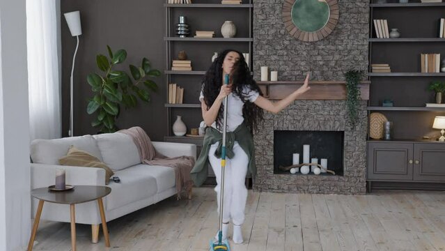 Happy young funny woman cleans in modern living room singing song using mop as microphone active energetic housewife enjoying cleaning washes floor ridiculous dancing cleaning agency advertisement