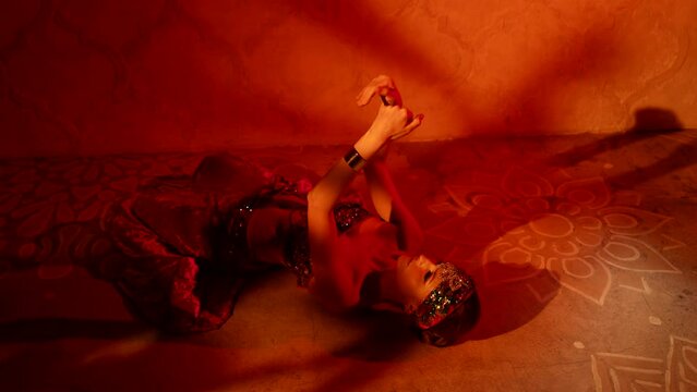 a woman with an adornment on head and in a sparkling bodice lies on the floor and moves hands beautifully against the wall with shadows. red light. the camera is moving
