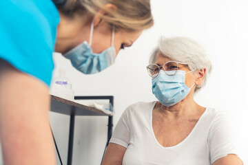 Fototapeta premium Senior lady with grey hair and her glasses on smiling sincerely at the doctor while wearing a face mask during a medical checkup. High-quality photo