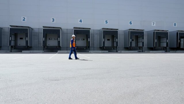 Engineer walks against large warehouse with open truck unloading gates. Specialist in uniform comes with inspection of large industrial building